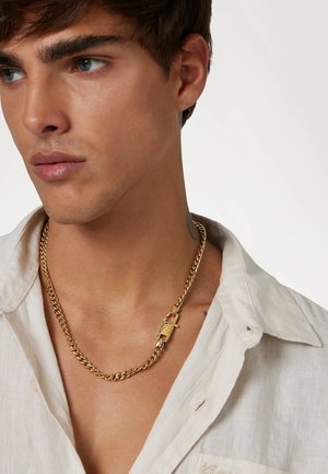 Gouden ketting met een gestructureerd schakeldesign en sluiting, gedragen over een lichtgekleurde shirt. Opvallend hardwaredetail op de sluiting.