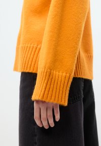 Πλεκτό πορτοκαλί πουλόβερ με ριμπ μανσέτες και τελείωμα. Απαλή υφή, εφαρμοστή γραμμή. Σκουρόχρωμο τζιν παντελόνι ορατό στο κάτω μέρος, χέρι ακουμπισμένο στο πλάι.