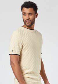 Suéter tejido de punto texturizado en color beige con un patrón de rejilla, cuello redondo y mangas cortas, que presenta puños y dobladillo acanalados en un tono azul marino oscuro.