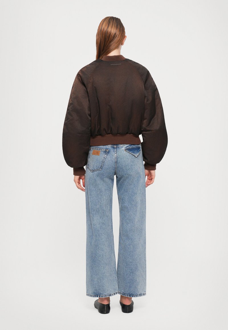 Veste bomber brune semi-transparente avec des manches bouffantes et des poignets côtelés, associée à un jean en denim bleu à jambes larges avec une poche plate.