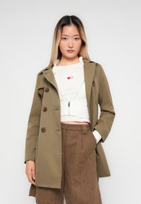 Junge Frau in einem beigen Trenchcoat, weißem, knappen Reißverschluss-Pullover mit zwei Herziconen und braunen Herringbone-Musterhosen, die steht.
