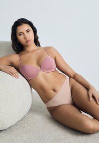 Lys rosa polstret bh med justerbare stropper og sømløst design, kombinert med nude høyhulede bikiniunderbukser på en myk beigemelert overflate.