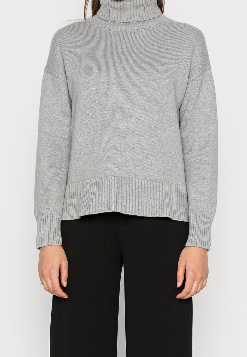 Femme portant un pull à col roulé côtelé gris clair à manches longues et un pantalon noir, debout devant un fond uni.