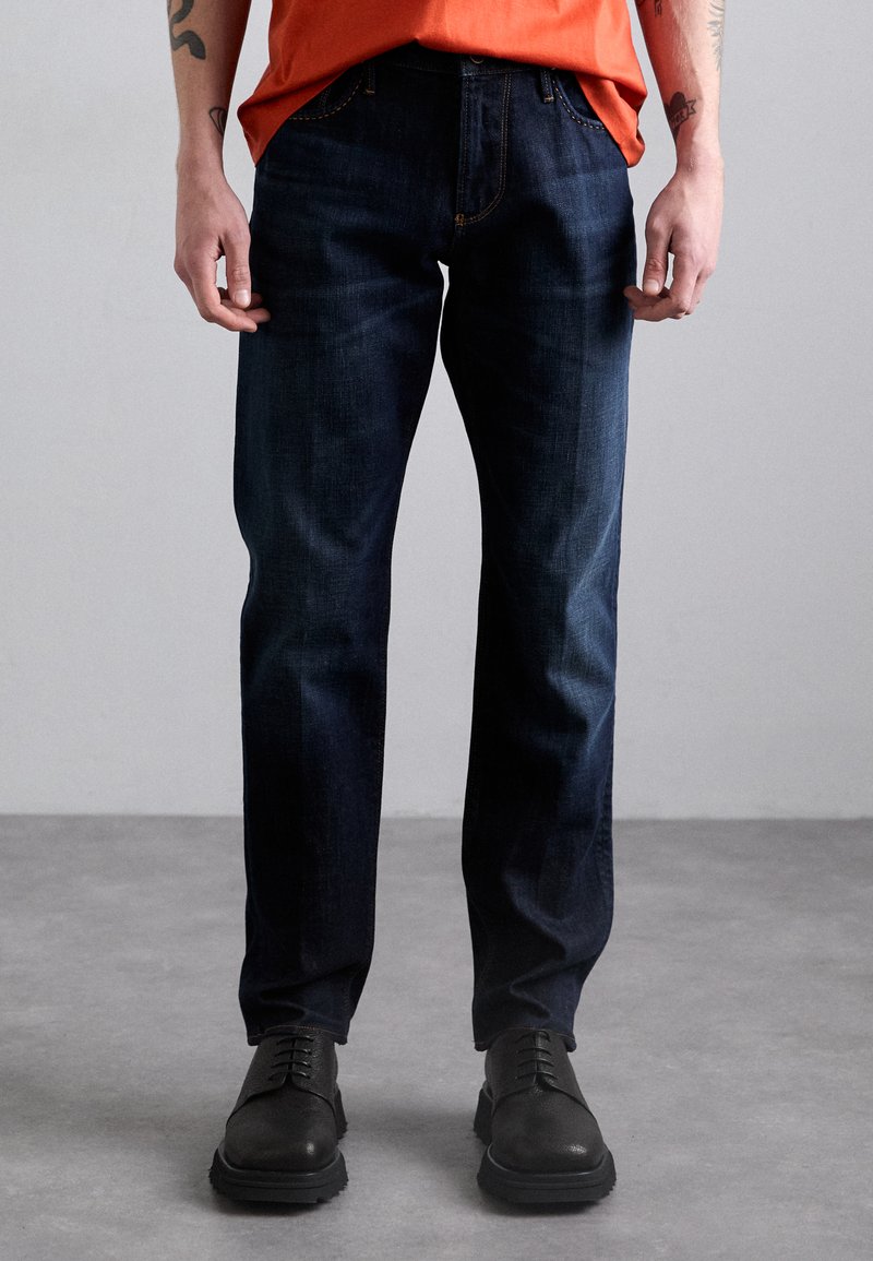 Dark blue denim jeans with a straight-leg design, featuring orange stitching and belt loops, paired with black leather shoes.