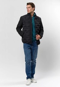 Mann iført svart vattert jakke over teal glidelåsgenser, blå jeans og hvite joggesko, stående med hendene i lommene og ser mot venstre.