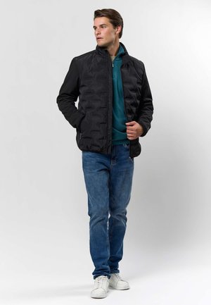 Homme portant une veste matelassée noire par-dessus un pull zippé bleu canard, jean bleu et baskets blanches, debout les mains dans les poches et regardant vers la gauche.