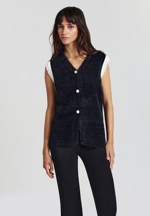 Donna con lunghi capelli scuri indossa un gilet nero peloso senza maniche con tre bottoni di perla sopra una maglietta bianca e pantaloni neri, su uno sfondo semplice.