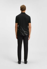 Zwarte polo met korte mouwen, versierd met witte geometrische lijnen op de achterzijde. Gecombineerd met zwarte broek en zwarte schoenen. Gladde textuur.