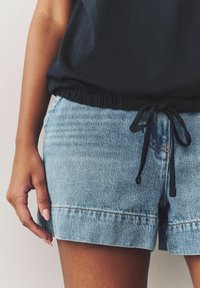 Heledad sinine denimsärg, millel on paelaga vöökoht ja veidi luitunud äär, kombineeritud musta t-särgiga. Suurendatud vaade, mis näitab käe detaile.