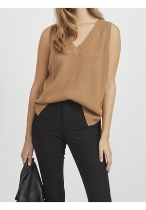 Femme portant un haut tricoté marron sans manches à col en V, rentré dans un pantalon noir taille haute, tenant une veste en cuir noire.