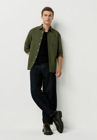 Cămașă căptușită verde măsliniu peste un tricou negru, asociată cu blugi închiși și cizme de culoare marron, rezistente. Cămașa are un buzunar pe piept și mânecile suflecate.