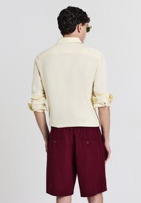 Chemise boutonnée jaune clair avec les manches retroussées, associée à un short bordeaux avec deux poches arrière et une coupe décontractée.