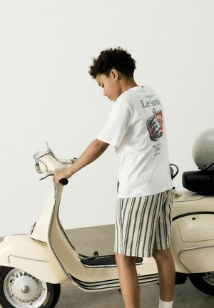 Enfant portant un t-shirt graphique blanc et un short beige rayé, interagissant avec un scooter vintage crème aux détails chromés.