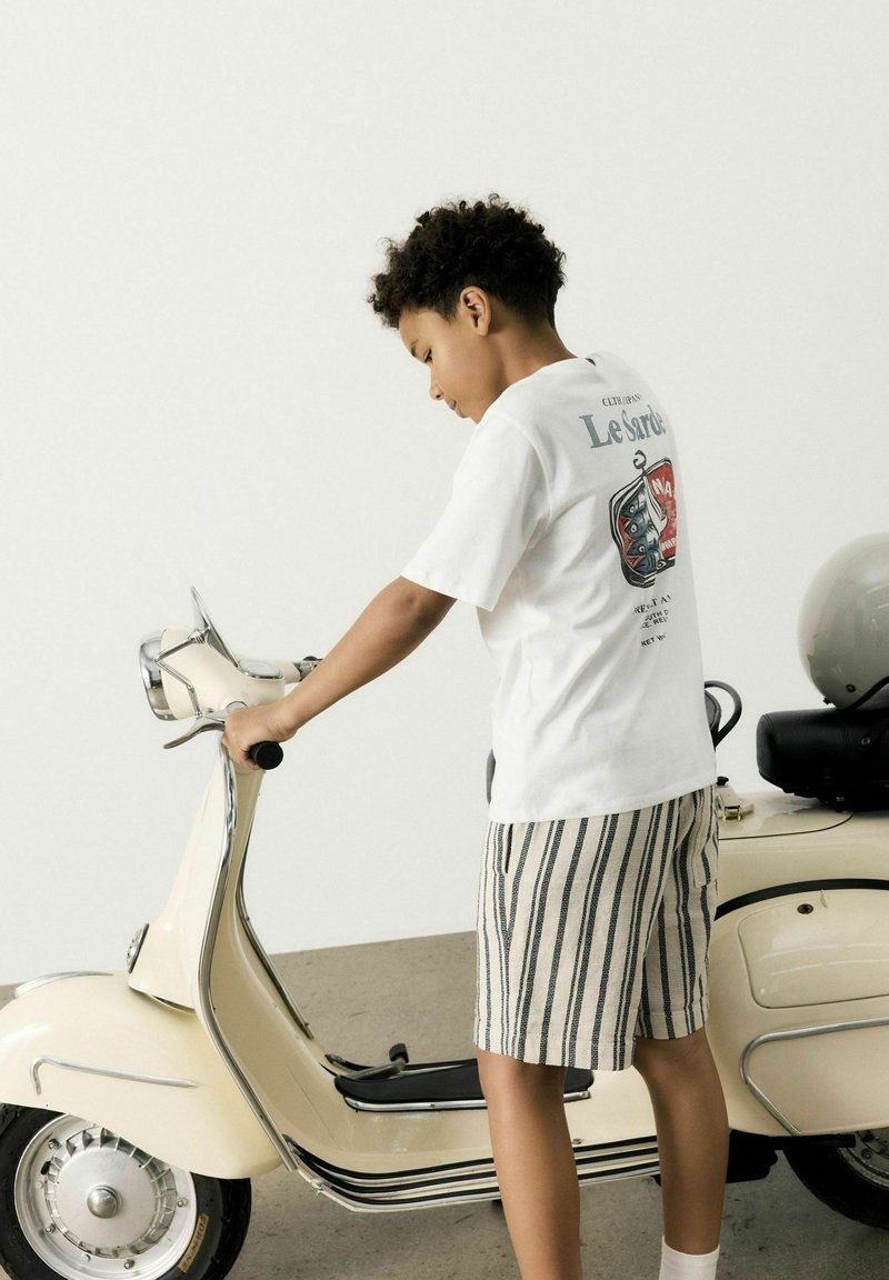 Dziecko w białej koszulce z grafiką i beżowych szortach w paski, bawiące się zabytkowym kremowym skuterem z chromowanymi detalami.