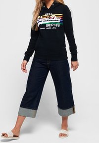 Donna che indossa un hoodie nero di SuperDry con strisce arcobaleno, jeans di denim scuri larghi e risvoltati, e ciabatte bianche, in piedi con le gambe divaricate.