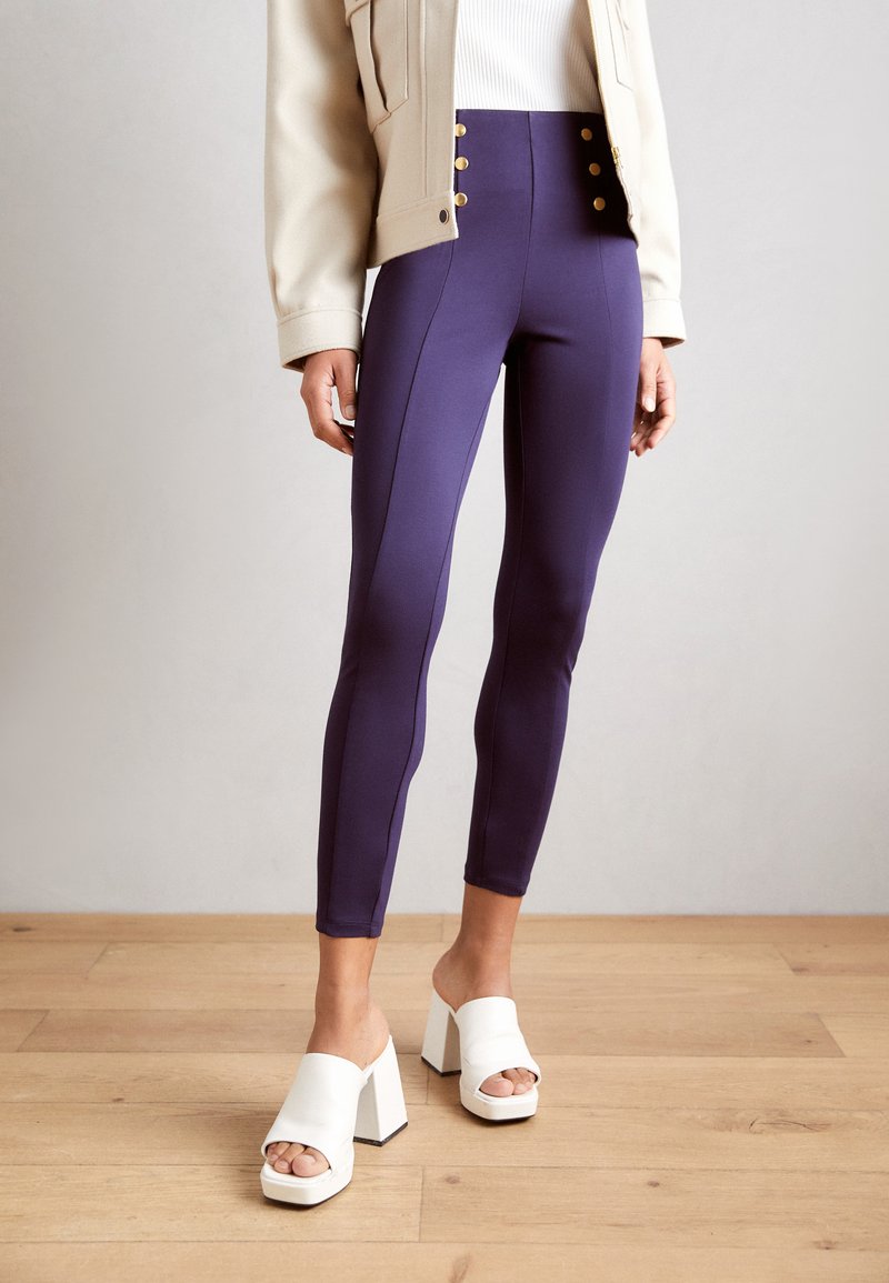 Hooglopende paarse leggings met zijknaden, gouden knopenaccenten, gecombineerd met witte open teen block-heel muilen op een houten vloer.
