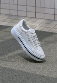 Witte sneaker met een glad leren bovenwerk en suède accenten, geperforeerde neus, platte veters en een gestructureerde rubberen zool met een zwarte streep.