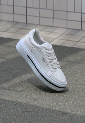 White sneaker with a smooth leather body and suede accents, perforated toe, flat laces, and a textured rubber sole with a black stripe.