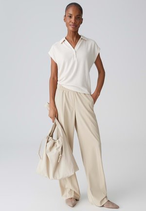 Vrouw die een wit hemd met korte mouwen en kraag draagt en beige broek met wijde pijpen, houdt een grote beige koordtas vast, staand tegen een effen achtergrond.