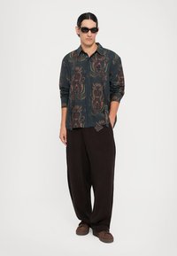 Long-sleeve button-up shirt in dark blue with intricate red and green floral patterns, paired with loose-fitting brown pants and brown shoes.