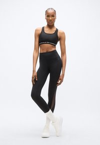 Μαύρο αθλητικό crop top με λογότυπο, μαύρο ψηλόμεσο κολάν με διαφανείς ενθέσεις, σετ με λευκά αθλητικά παπούτσια και κάλτσες.