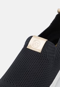Schwarze Strick-Slip-On-Sneaker mit weißer Sohle und goldenem Riemen, der ein geprägtes, kreisförmiges Logo an der oberen Vorderseite zeigt.