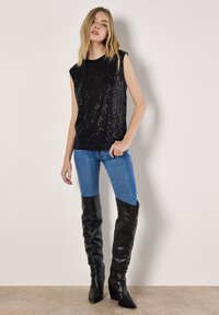 Jonge vrouw met een mouwloze zwarte top met pailletten, blauwe strakke jeans en zwarte leren laarzen tot over de knie, staand tegen een effen witte muur.