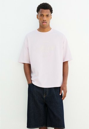 Jeune homme aux cheveux courts et bouclés portant un t-shirt oversize rose clair et un short en jean foncé, debout contre un mur blanc.