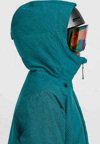 Teal ski-jack met een gestructureerde afwerking, een aansluitende capuchon en een ritssluiting aan de voorkant. Gecombineerd met een zwarte helm en spiegeltrotters.