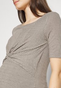 Gravid kvinna som bär en ribbad, beige, kortärmad topp med en vriden knutdetalj under bysten.