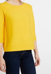 Gelbes, leichtes Strickoberteil mit Dreiviertelärmel, Rundhalsausschnitt und geripptem Saum. Tragen mit dunklen Jeans. Glatte Textur.