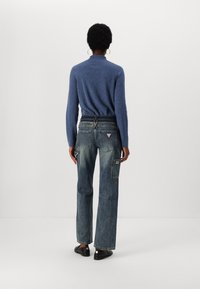 Blue knit sweater with a high collar, paired with loose-fitting denim jeans featuring pockets and a patch on the back. Black shoes complete the outfit.