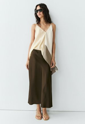Femme aux cheveux ondulés foncés portant des lunettes de soleil noires, un haut asymétrique crème et gris, une jupe marron foncé et des sandales beige, debout contre un mur blanc.