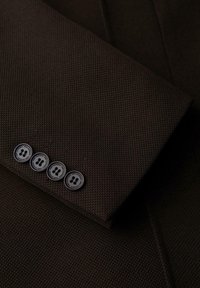 Tejido marrón texturizado con un aspecto a medida. Cuatro botones negros en el puño, con detalles de diseño circular, acentúan la manga.