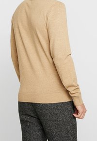 Personne portant un pull à manches longues beige et un pantalon texturé gris foncé, vue de dos sur un fond clair uni.