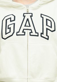 Λευκό φούτερ με φερμουάρ, με μεγάλα γράμματα με μαύρο περίγραμμα που σχηματίζουν τη λέξη "GAP" στο στήθος και τσέπη καγκουρό μπροστά.