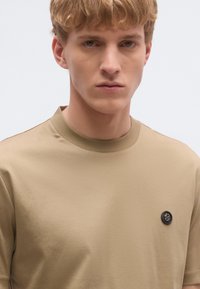 Bézs színű rövid ujjú póló, kerek nyakkivágással és bal oldalon fekete gombdísszel. Simább, puha tapintású anyagból készült.