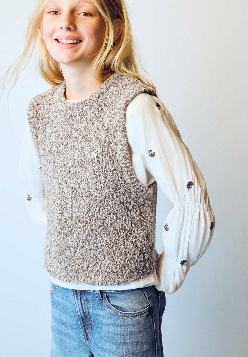 Jeune fille souriante aux longs cheveux blonds portant un gilet beige en tricot par-dessus un chemisier blanc avec une petite broderie fleurie et un jean bleu.