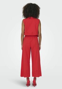 Rode tweedelige outfit met een mouwloos topje en brede broek met oogjesdetails aan de zoom. Materiaal lijkt lichtgewicht en gestructureerd.