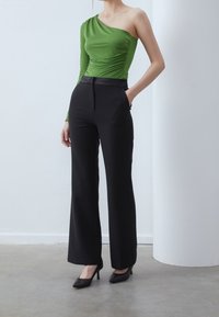 Kvinna klädd i en grön enärmad topp och svarta vida byxor med svarta högklackade skor, stående med en hand i fickan.