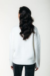 Hvit, strikket genser med avslappet passform, ribbestrikket kant og senkede skuldre, kombinert med mørke, teksturerte jeans. Modellens hår faller ned.