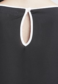 Vêtement noir avec bordure blanche au niveau de l'encolure, comportant une petite ouverture en forme de trou de serrure fermée par un bouton blanc en haut.