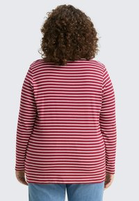 Persoon met krullend haar, gekleed in een rood-wit gestreepte lange mouwen shirt en blauwe spijkerbroek, met de rug naar ons gekeerd tegen een effen achtergrond.