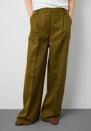 Person trägt olivgrüne Hose mit weitem Bein, vorne mit Falten und Taschen, kombiniert mit braunen, gewebten Schuhen mit spitzen Zehen.