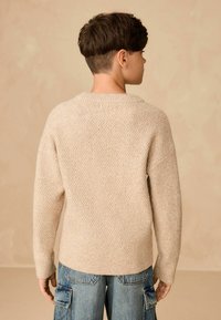 Beżowy sweter dzianinowy o teksturowanym wzorze, z szerokim dekoltem i ściągaczami, noszony z niebieskimi spodniami cargo z bocznymi kieszeniami.