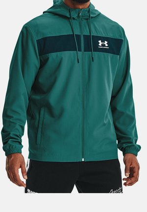 Mężczyzna w turkusowej bluzie z kapturem na suwak z czarnym paskiem i logo Under Armour na piersi, w połączeniu z czarnymi spodenkami sportowymi.
