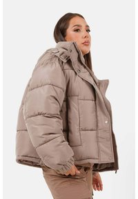 Vrouw in een oversized taupe pufferjack met hoge kraag en drukknoopsluiting, gecombineerd met bijpassende broek, staande zijwaarts op een witte achtergrond.