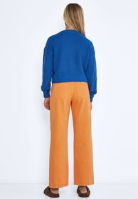 Blå, ribbade tröja i kombination med högmidjade, vida orange byxor. Bakifrån syns platta fickor och en avslappnad passform. Bruna, texturerade skor.