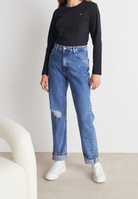 Hoge taille blauwe spijkerbroek met een versleten knie, opgerolde boorden en een klassiek vijf-pocket ontwerp, gecombineerd met een zwart shirt met lange mouwen.