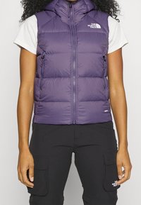 Fioletowa kamizelka puchowa z wysokim kołnierzem, zamkiem błyskawicznym z przodu i dwoma bocznymi kieszeniami. Materiał jest gładki i ocieplany. Logo na lewej piersi.